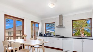 A modern kitchen with white cabinets, a wooden dining table, and a large window. - Kozyguru | Thornleigh | Townhouses (Thornleigh)