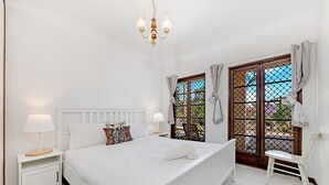 A white bedroom with a bed, bedside tables, a chandelier, and a window with curtains. - Kozyguru | Thornleigh | Townhouses (Thornleigh)