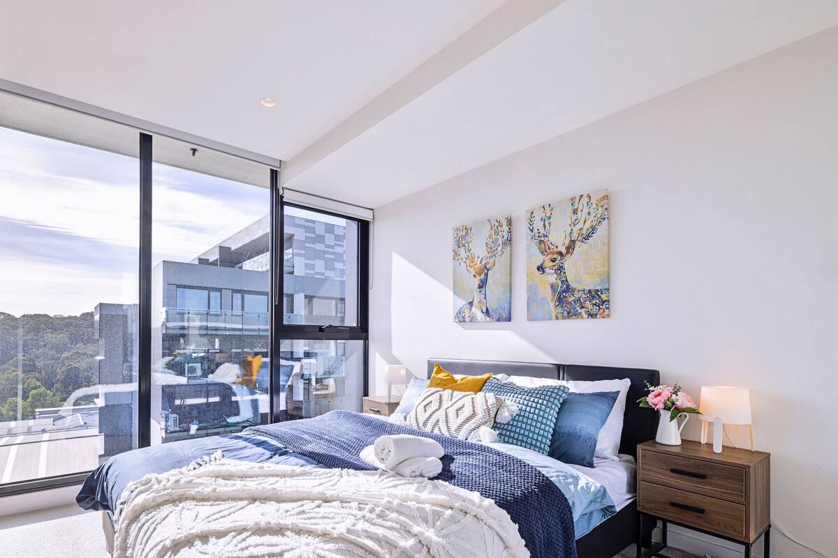 A modern bedroom with a large window, a bed with multiple pillows, a nightstand, and two wall-mounted paintings.