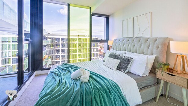 Un dormitorio moderno con una cama grande, una mesita de noche y vistas de la ciudad a través de amplias ventanas.