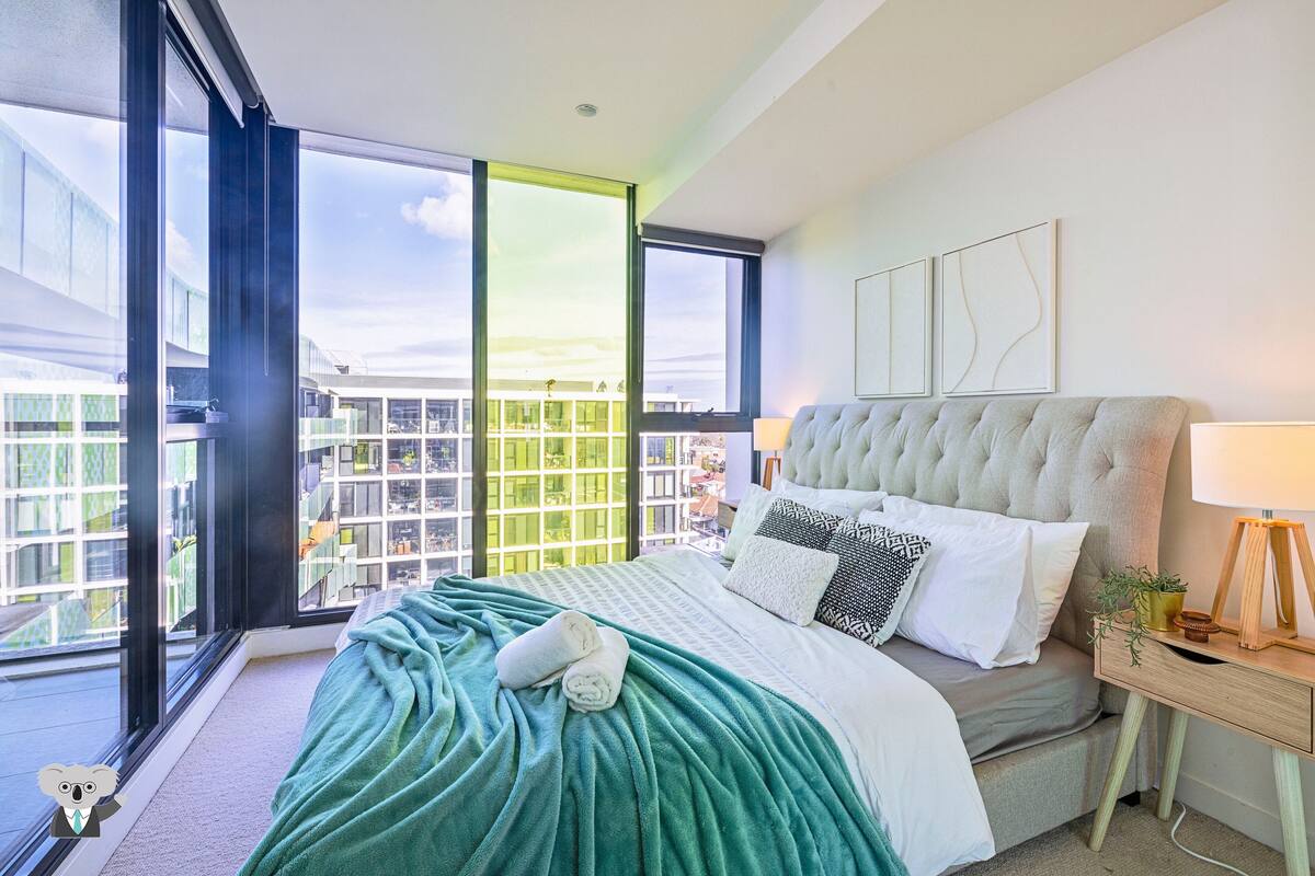 A modern bedroom with a large bed, a nightstand, and a view of the city through large windows.