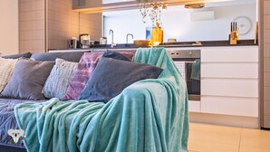 Una sala de estar moderna con un sofá cubierto con una manta color turquesa, y un área de cocina al fondo con un lavamanos y electrodomésticos sobre la encimera.