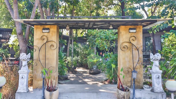 A garden pathway leading to a pergola with two columns, surrounded by potted plants and a small water feature.