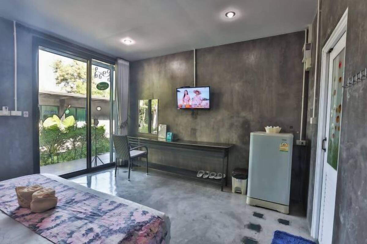 Una habitación de hotel moderna con un gran ventanal corredero a un balcón, un televisor de pantalla plana montado en la pared, un refrigerador pequeño y un banco con cojines.