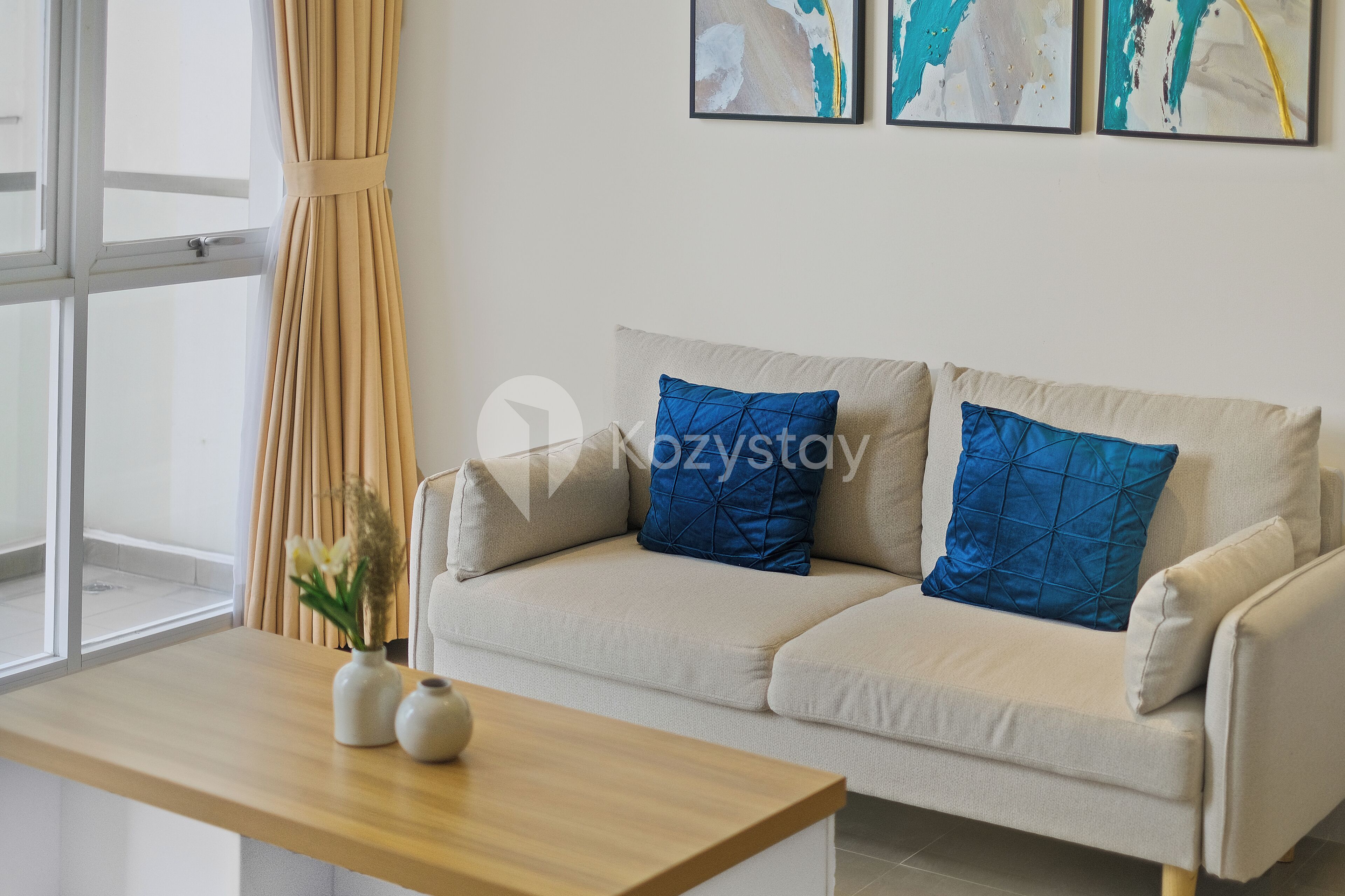 A modern living room with a beige sofa, two blue pillows, a wooden coffee table, and a vase with flowers. There are abstract paintings on the wall and a window with curtains.