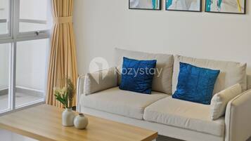 A modern living room with a beige sofa, two blue pillows, a wooden coffee table, and a vase with flowers. There are abstract paintings on the wall and a window with curtains.