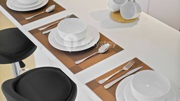 A dining table set with white plates, bowls, cutlery, and a stack of white cups.