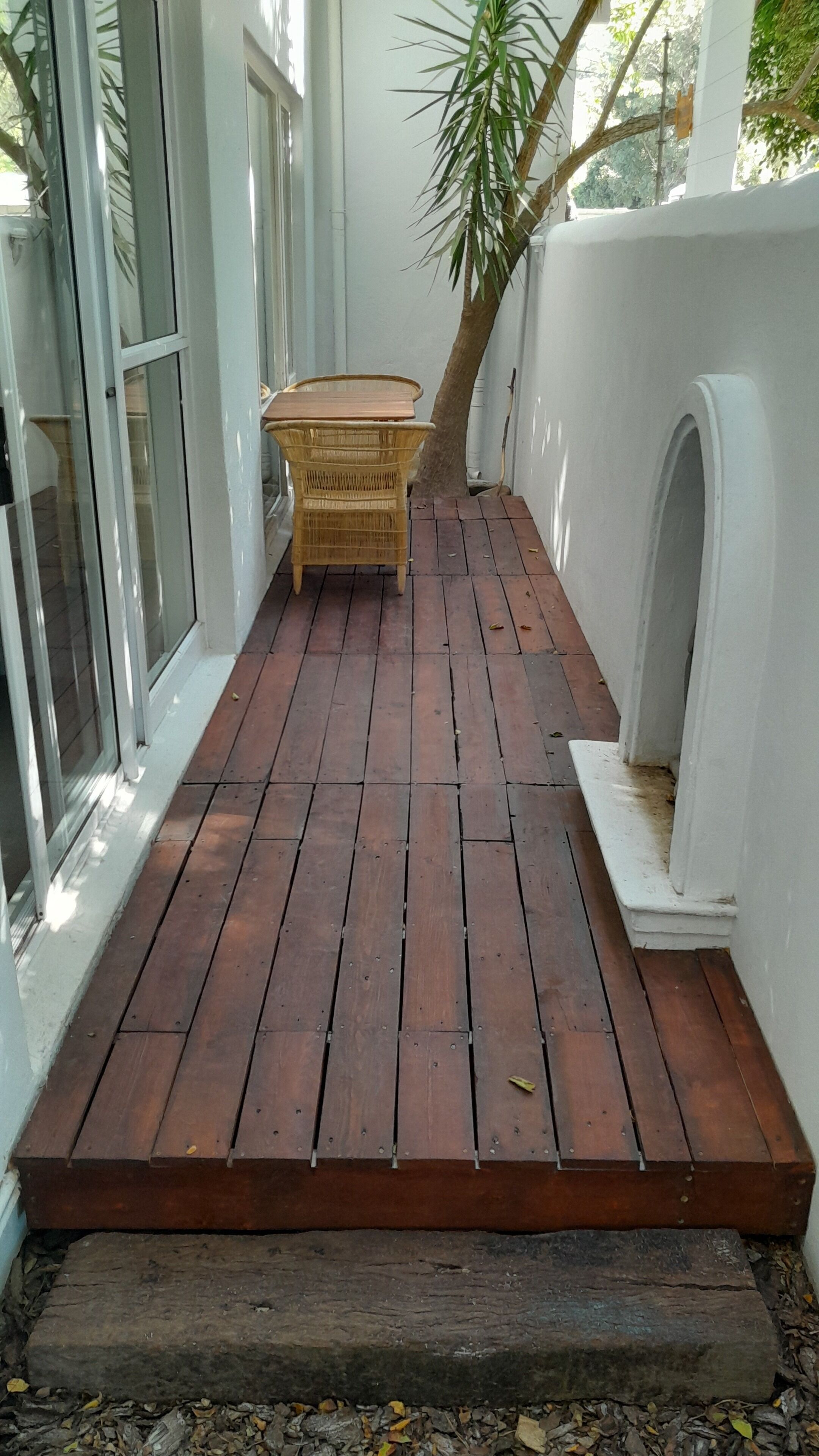 A wooden deck with a wicker table and chairs, surrounded by white walls and a tree.