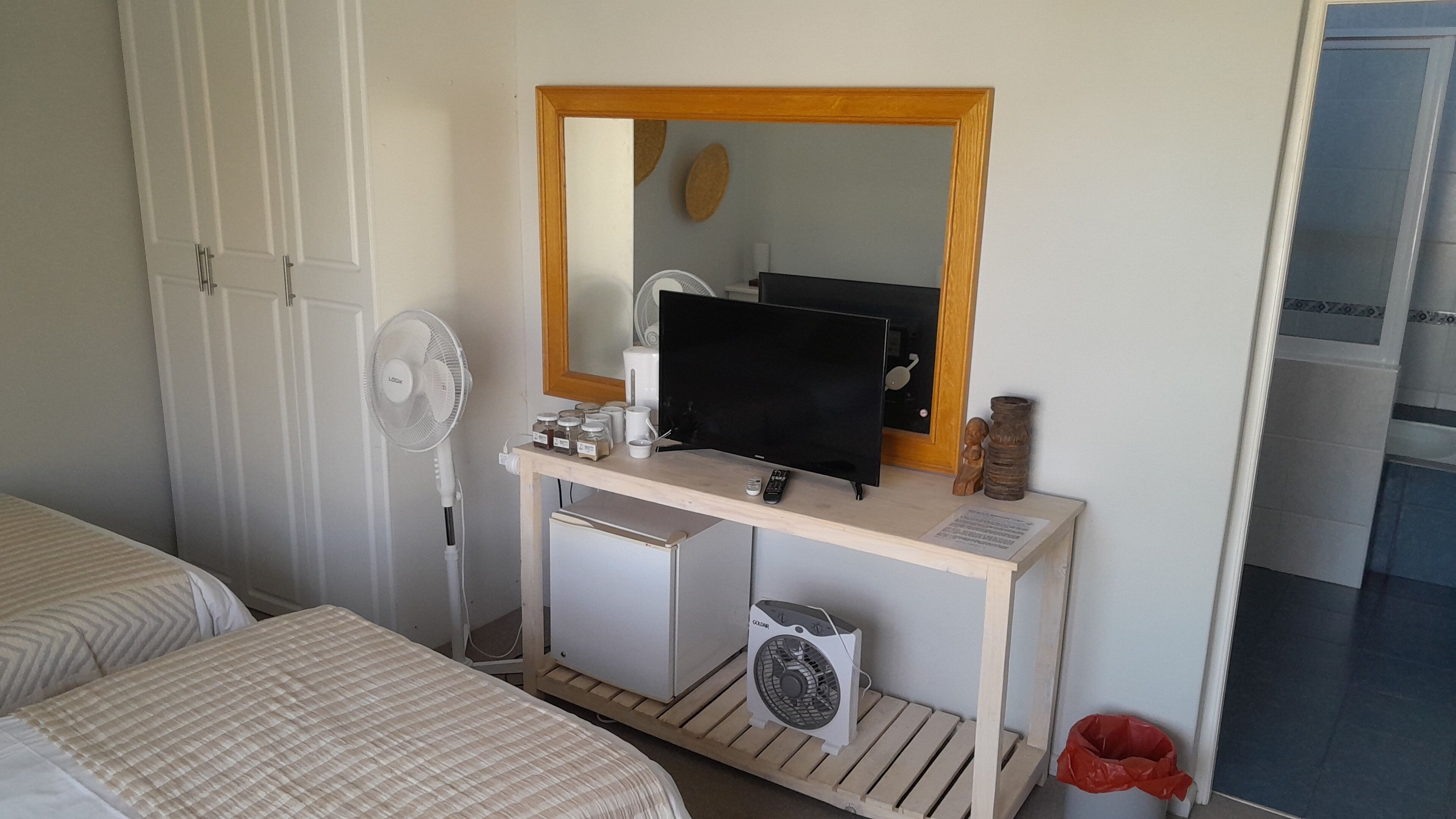 A bedroom with a bed, a desk with a computer, a mirror, a fan, and a trash bin.