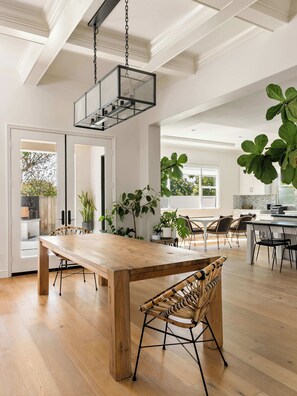Un vaste espace de salle à manger avec une table en bois et des chaises en osier, une cuisine dotée d’une grande fenêtre et un luminaire suspendu.
