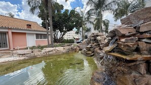 Una piscina con cascada, rodeada de muros de piedra y palmeras, con un edificio al fondo.