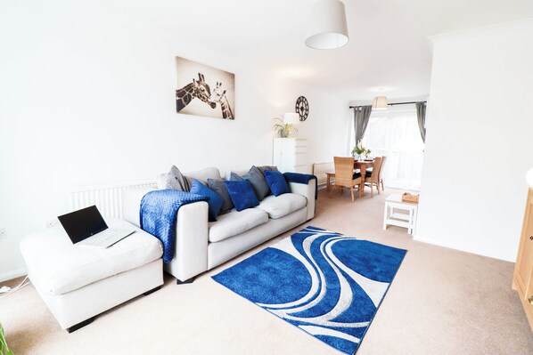 Ein modernes Wohnzimmer mit einem weißen Sofa, einem blauen Teppich und einem Laptop auf einem kleinen Hocker. Im Hintergrund befindet sich ein Essbereich mit Tisch und Stühlen.