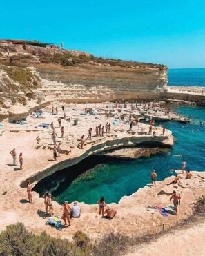 Une zone côtière avec une piscine naturelle entourée d’un paysage rocheux, où l’on peut observer des personnes pratiquant diverses activités.