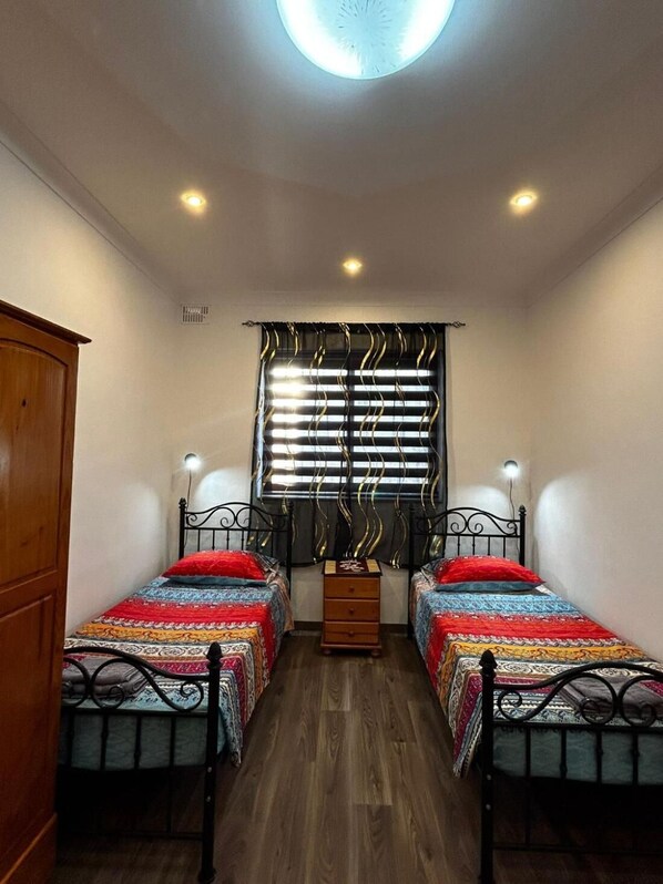 A small, well-lit room with two single beds, a wooden wardrobe, a window with curtains, and a ceiling light.