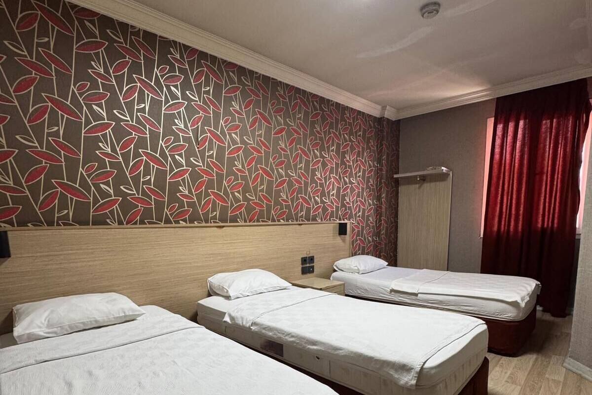 Habitación de hotel con tres camas individuales, papel tapiz estampado, cabecero de madera, una lámpara de techo y una cortina roja.
