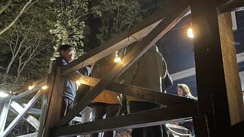 Une structure en bois dotée d’une ossature métallique, qui pourrait être une terrasse ou un balcon, agrémentée de guirlandes lumineuses et sur laquelle se tiennent des personnes.
