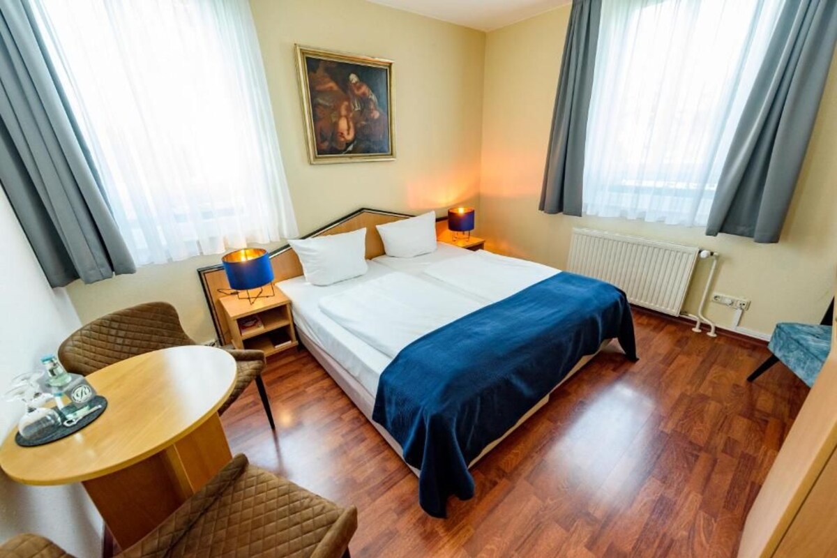A hotel room with a bed, a desk, a chair, a radiator, and a painting on the wall.