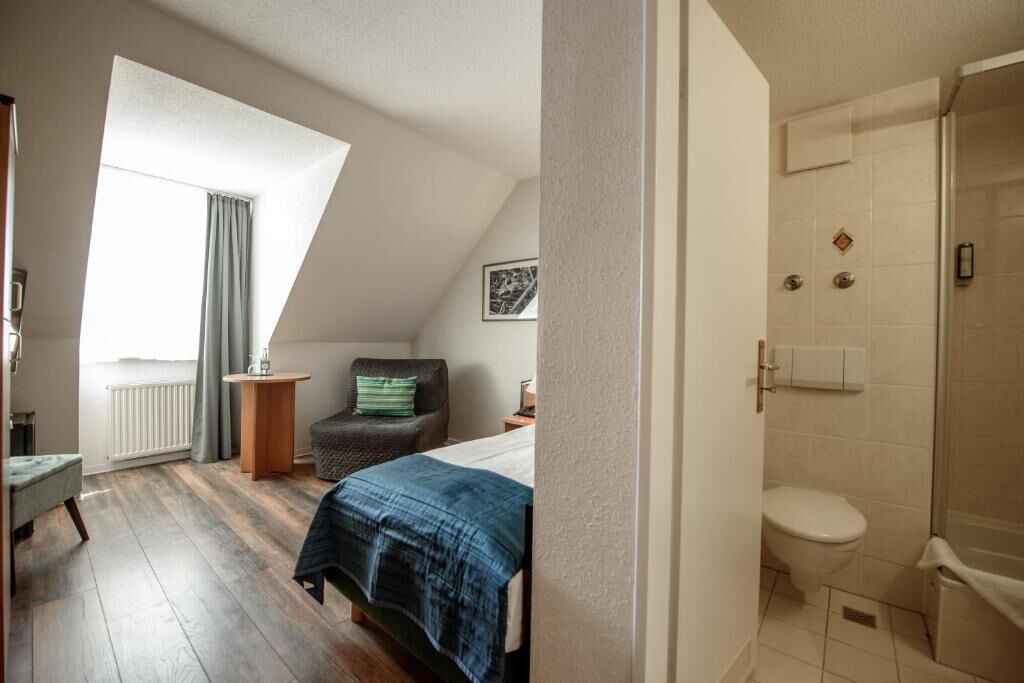 A bedroom with a bed, a chair, a small table, a radiator, and a window with curtains.