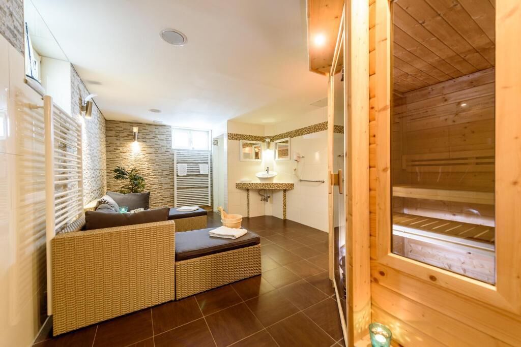 A modern spa room with a wooden sauna, a seating area with a sofa and cushions, a small sink, and a mirror.