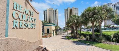 Un camino que lleva al Resort Beach Colony, con un letrero, palmeras y edificios altos al fondo.