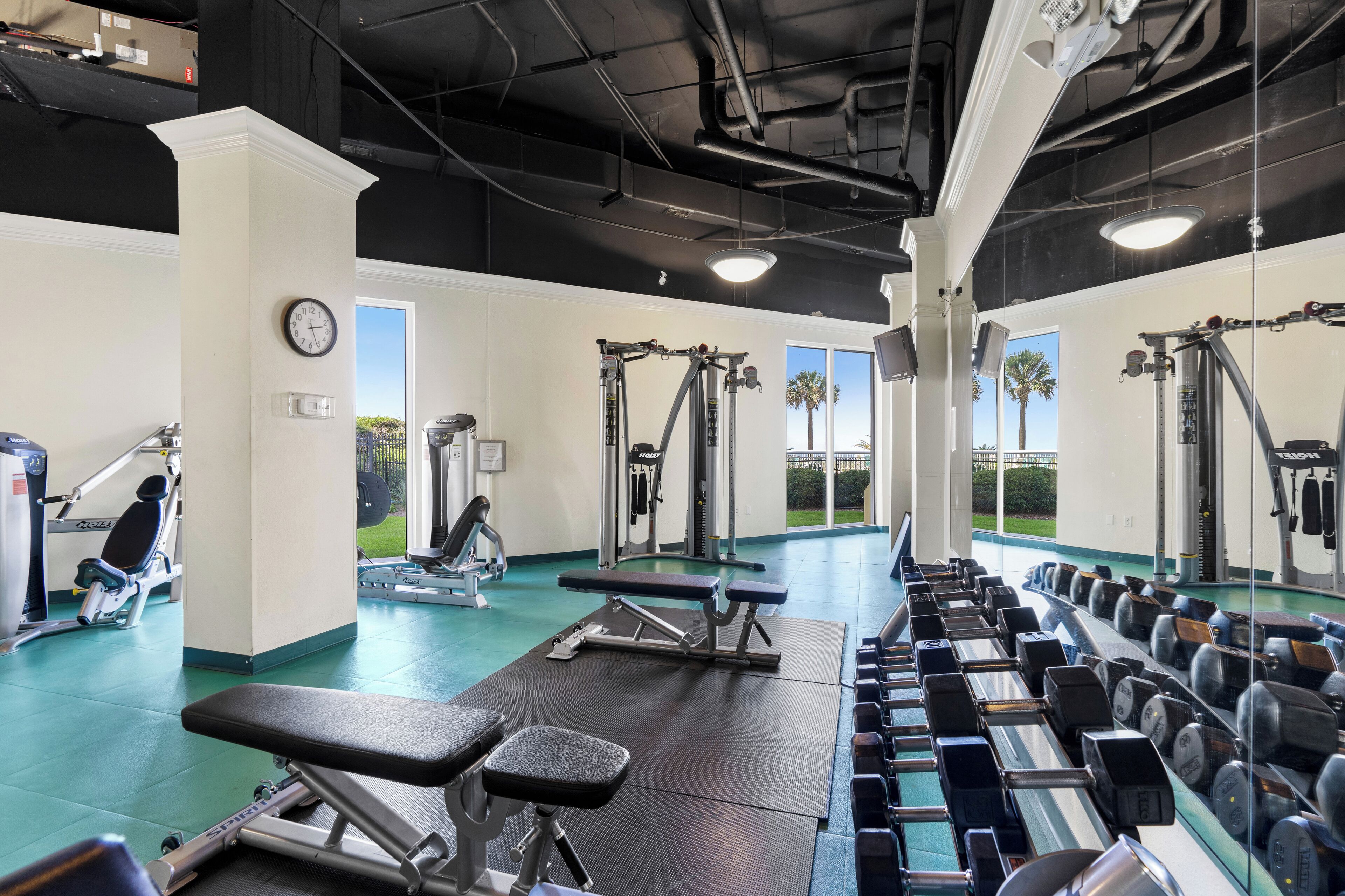 Une salle de sport équipée de divers appareils d’exercice, avec une horloge et une vue sur les palmiers à travers les fenêtres.