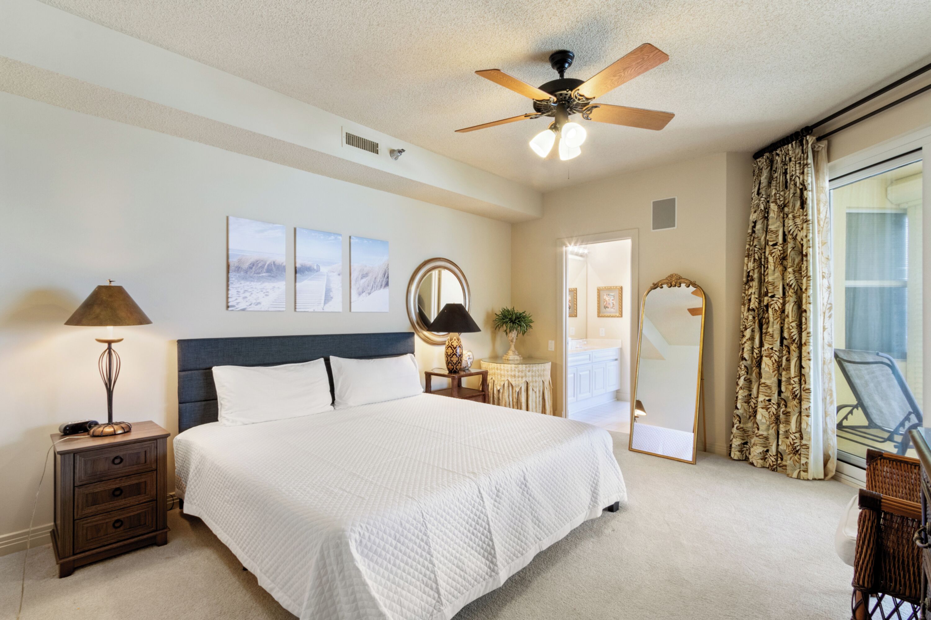 A bedroom with a large bed, a ceiling fan, a nightstand with a lamp, a mirror, and a view of the outside through a window with curtains.