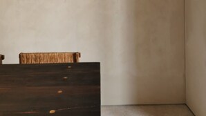Una habitación minimalista con una mesa de madera oscura y una silla tejida, una pared lisa y un suelo de hormigón.
