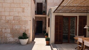 Un estrecho patio con muros de piedra, plantas en macetas, una mesa de madera y una puerta de vidrio que da a un balcón.