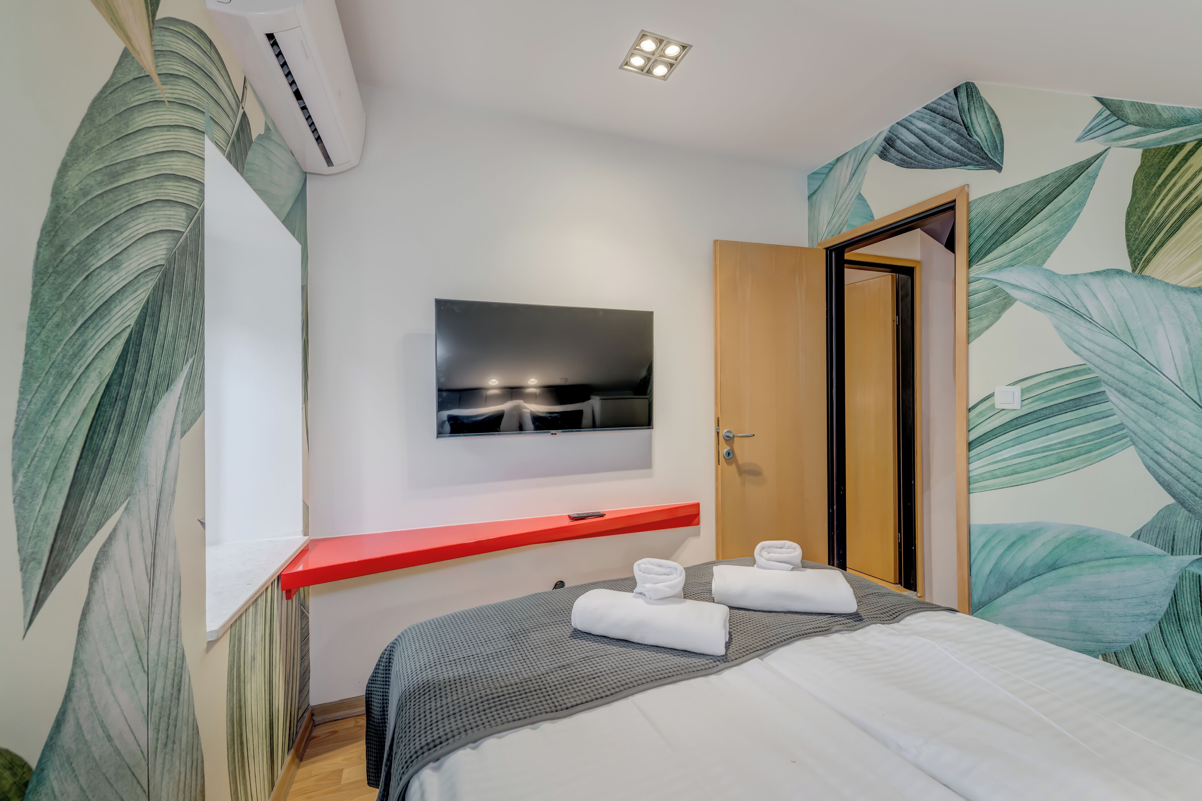 A hotel room with a bed, a TV mounted on the wall, a red shelf, and a door.