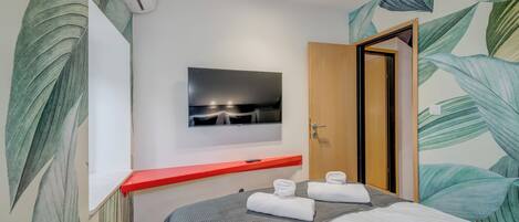 A hotel room with a bed, a TV mounted on the wall, a red shelf, and a door.