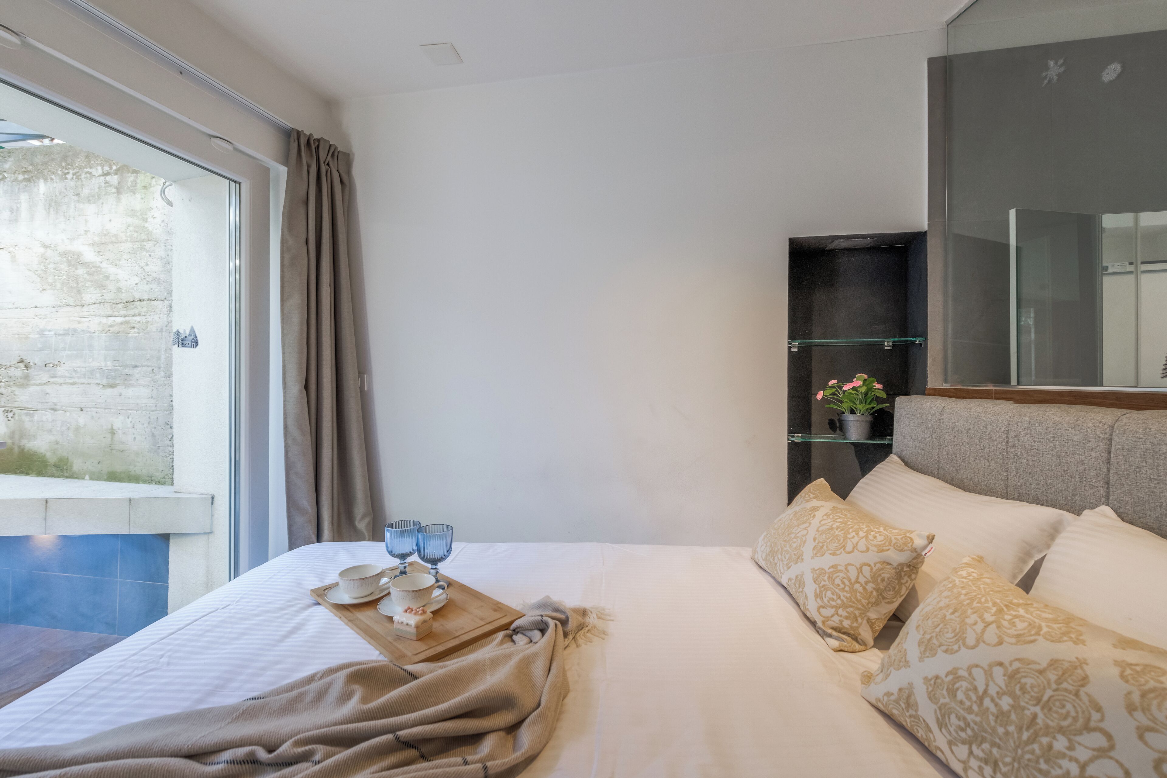 A hotel room with a bed, a bedside table with a tray of breakfast items, a window with curtains, a potted plant, and a mirror.