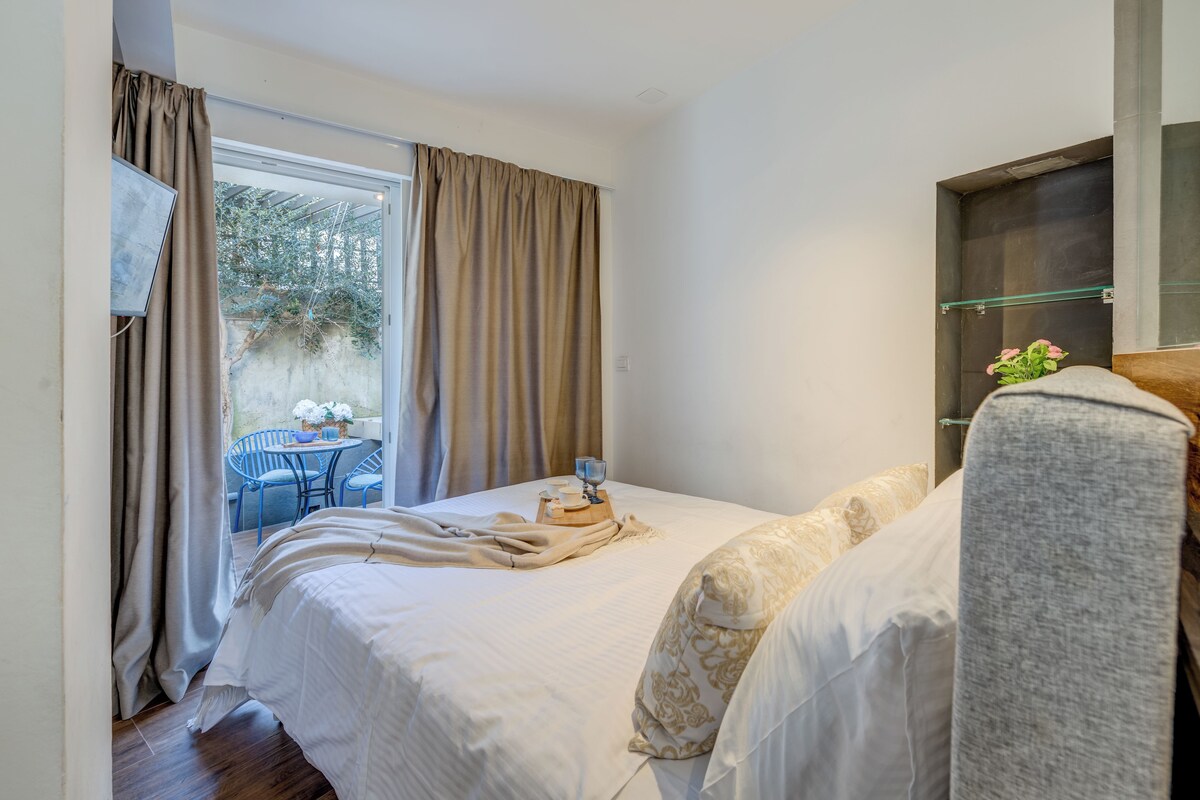 Une chambre à coucher comprenant un lit, une table de chevet, une fenêtre avec des rideaux, une chaise et une petite table sur laquelle est posé un vase.