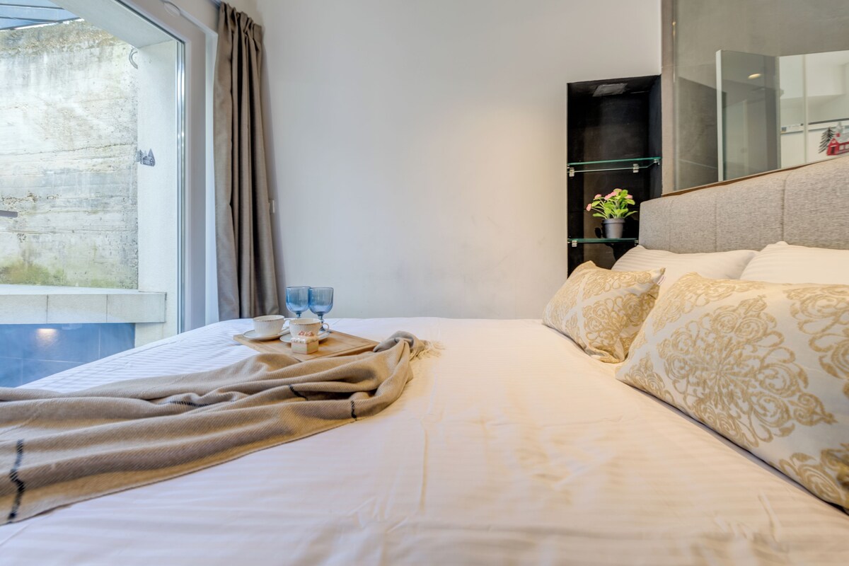 Habitación de hotel con una cama, una mesita de noche con vasos, una planta en maceta y vistas al exterior.