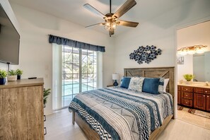 A bedroom with a large bed, a ceiling fan, a window with curtains, a wall art piece, and a bathroom with a vanity and mirror.