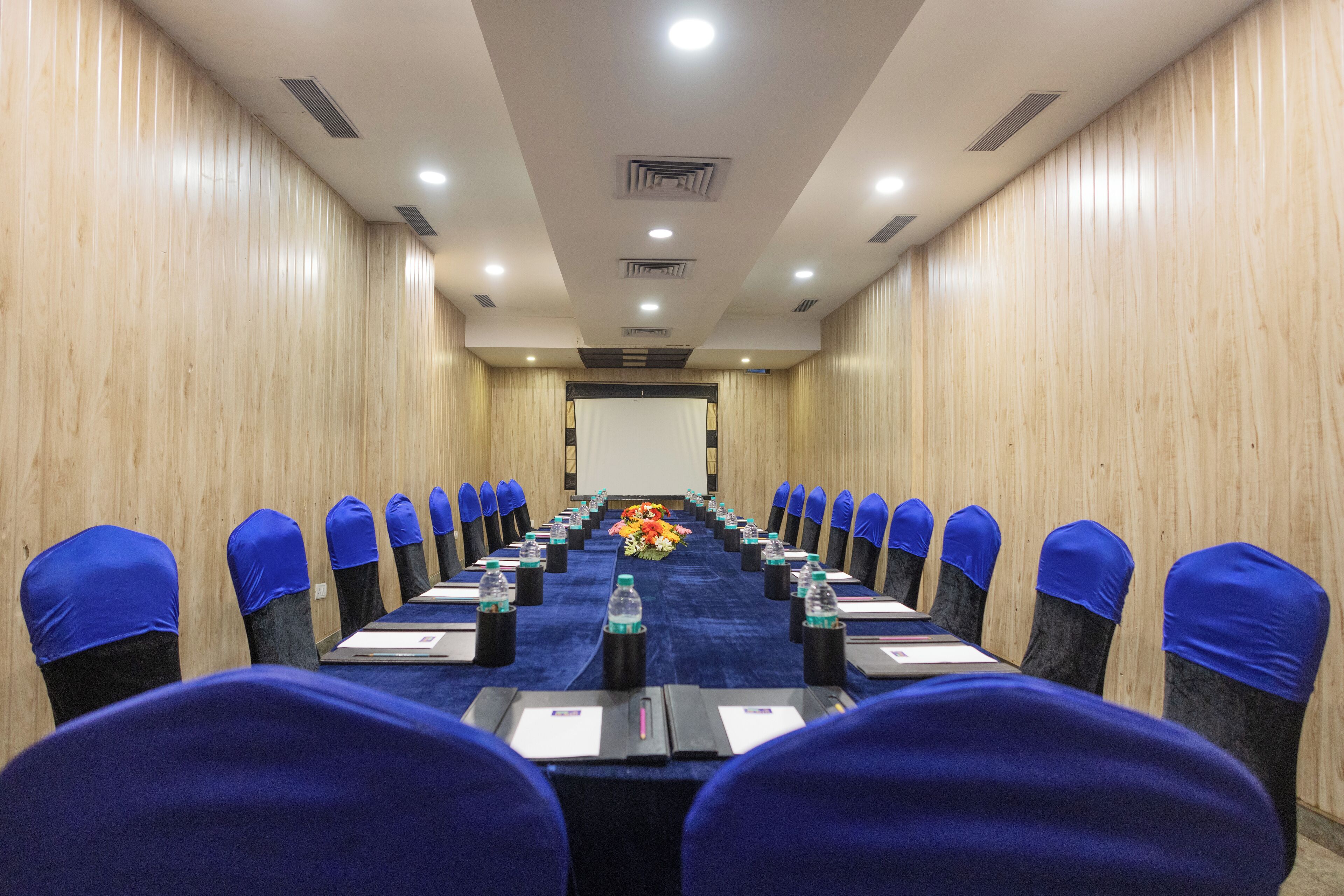 Une salle de conférence avec une longue table, des chaises bleues et un arrangement floral au centre.