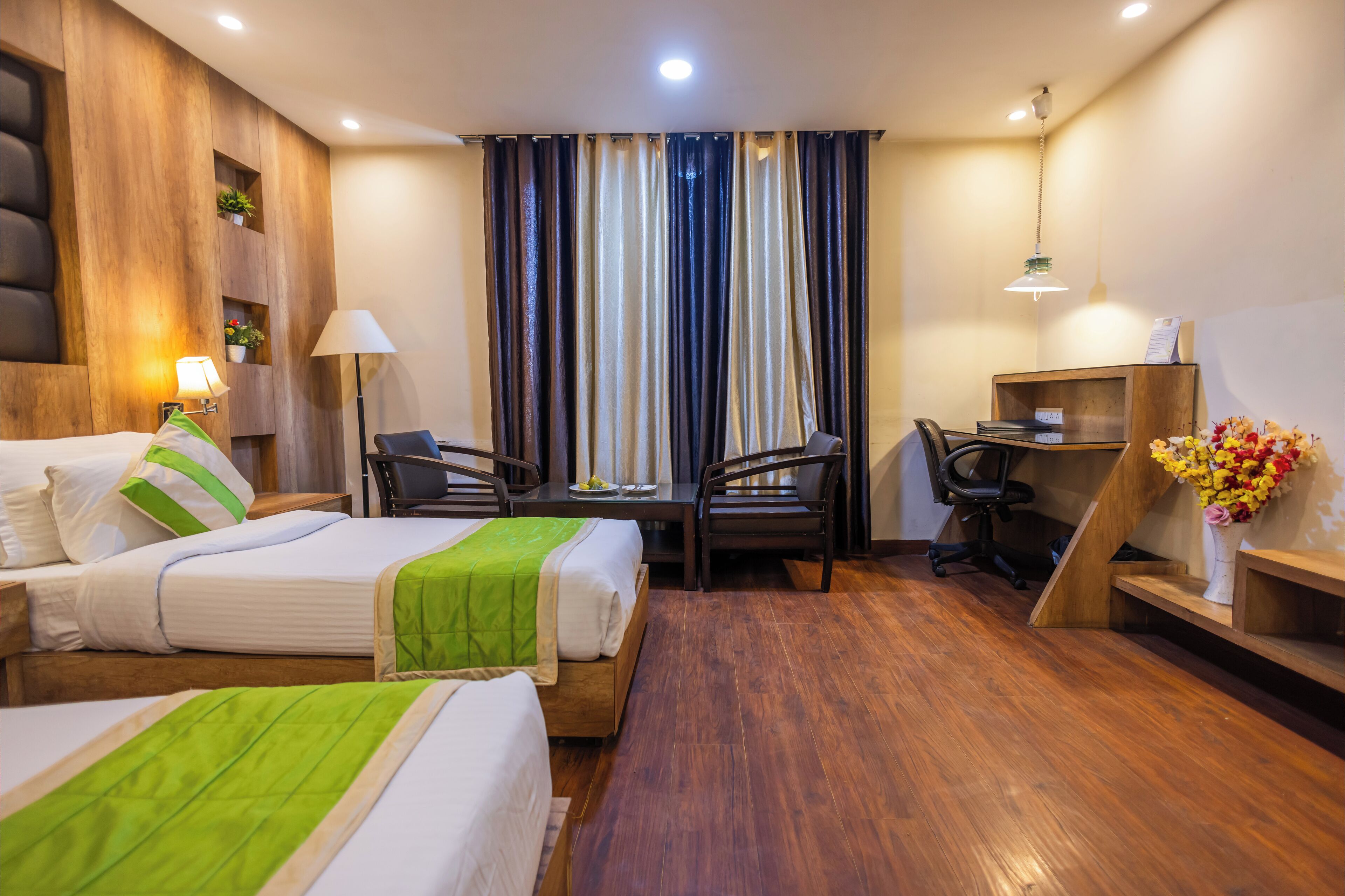 Habitación de hotel con dos camas, un escritorio, sillas, una lámpara y una estantería de madera con flores.