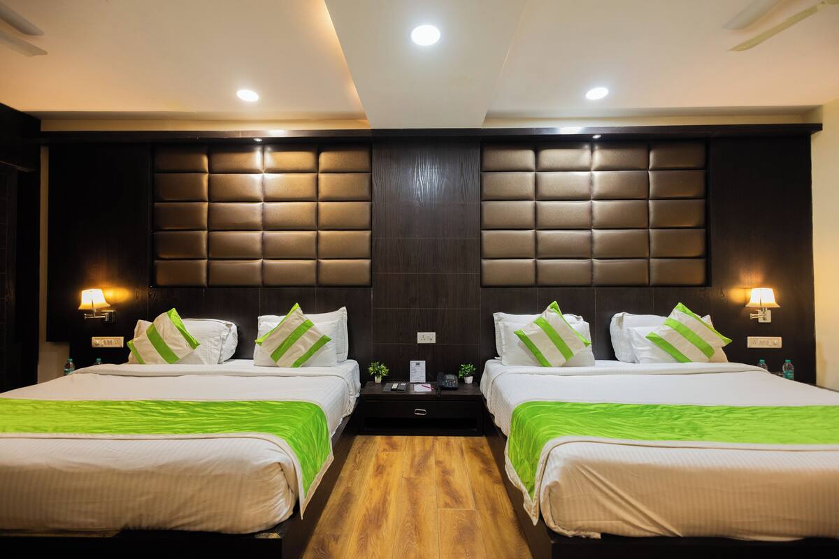 A hotel room with two beds, each with white bedding and green accents, a wooden floor, a dark headboard, and wall-mounted lamps.