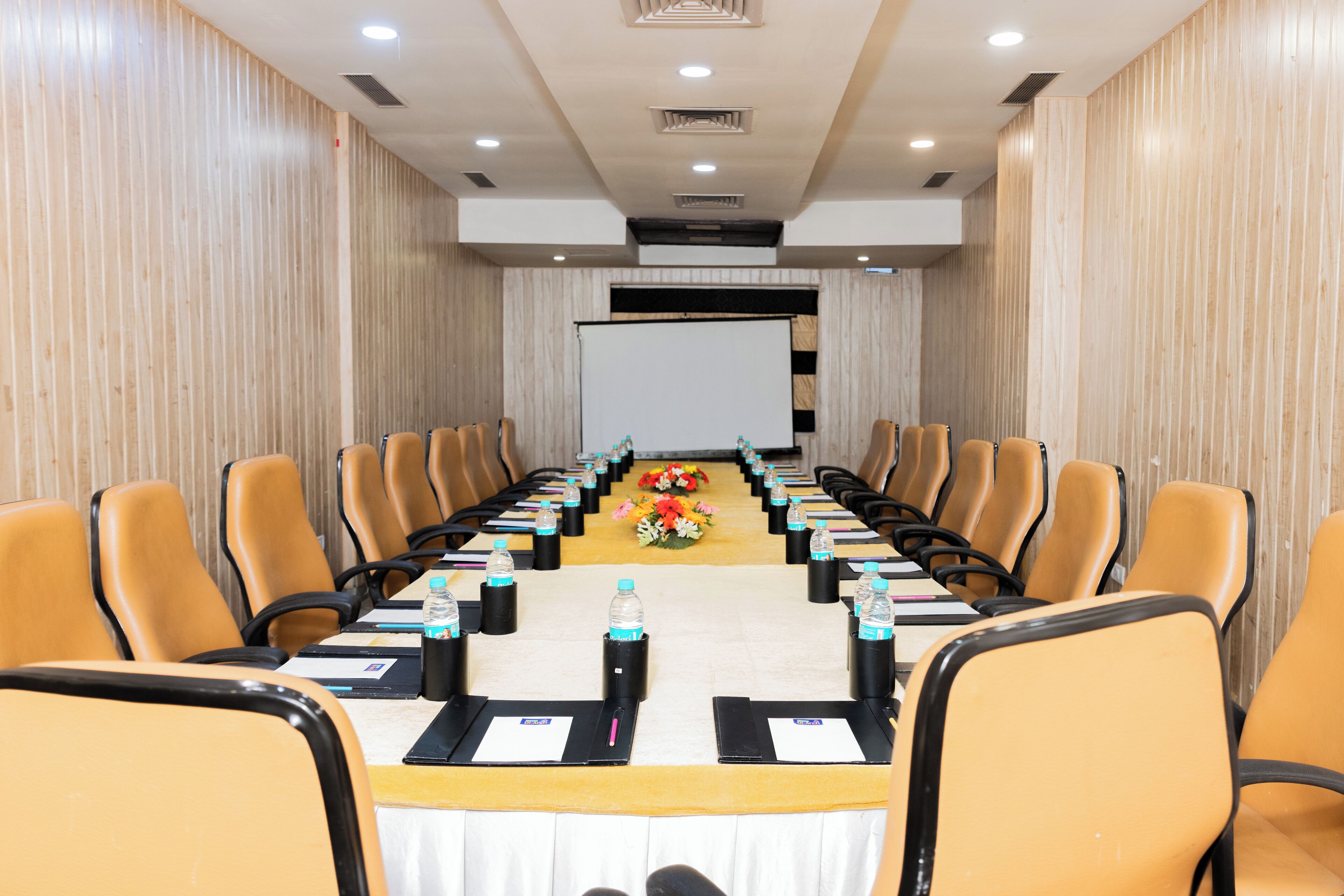 Un salón de conferencias con una larga mesa, sillas, botellas de agua y una pantalla de proyector.