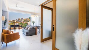 Ampla sala de estar com design moderno, com um sofá, uma poltrona e uma mesa de jantar. Uma grande porta deslizante dá acesso a um terraço ao ar livre com vista para uma encosta.