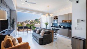 Uma sala de estar moderna com um sofá, uma mesa de centro e uma TV. Há uma área de cozinha com uma mesa de jantar e cadeiras. O quarto possui uma janela grande com vista para um horizonte urbano.
