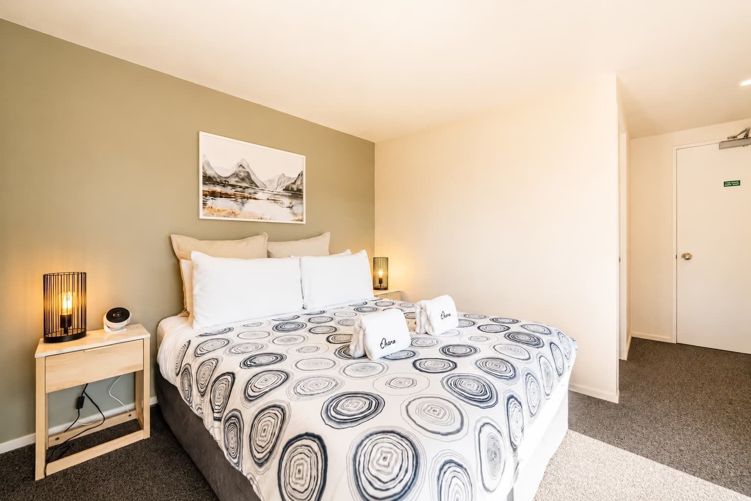 A hotel room with a bed, a nightstand, a lamp, a painting, and a door.