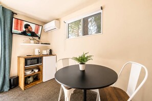 Ein kleiner Raum mit rundem Tisch, weißen Stühlen, einem kleinen Topfpflanzen und einem an der Wand befestigten Fernseher, sowie einem Fenster mit Blick auf Bäume.