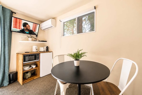 A compact room with a round table, white chairs, a small potted plant, a television mounted on the wall, and a window with a view of trees.