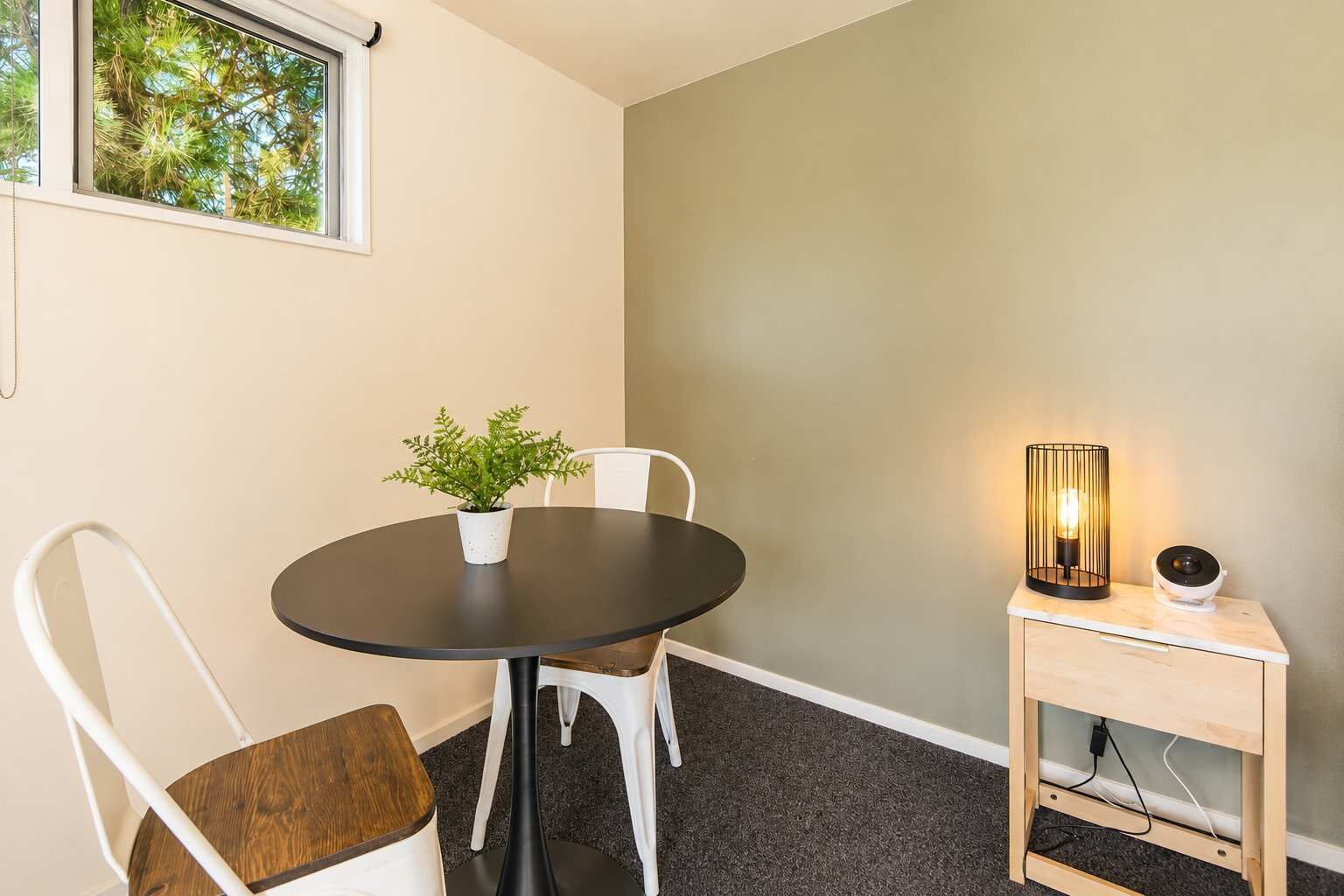 A small dining area with a round table, two white chairs, a potted plant, a lamp, and a wooden side table.