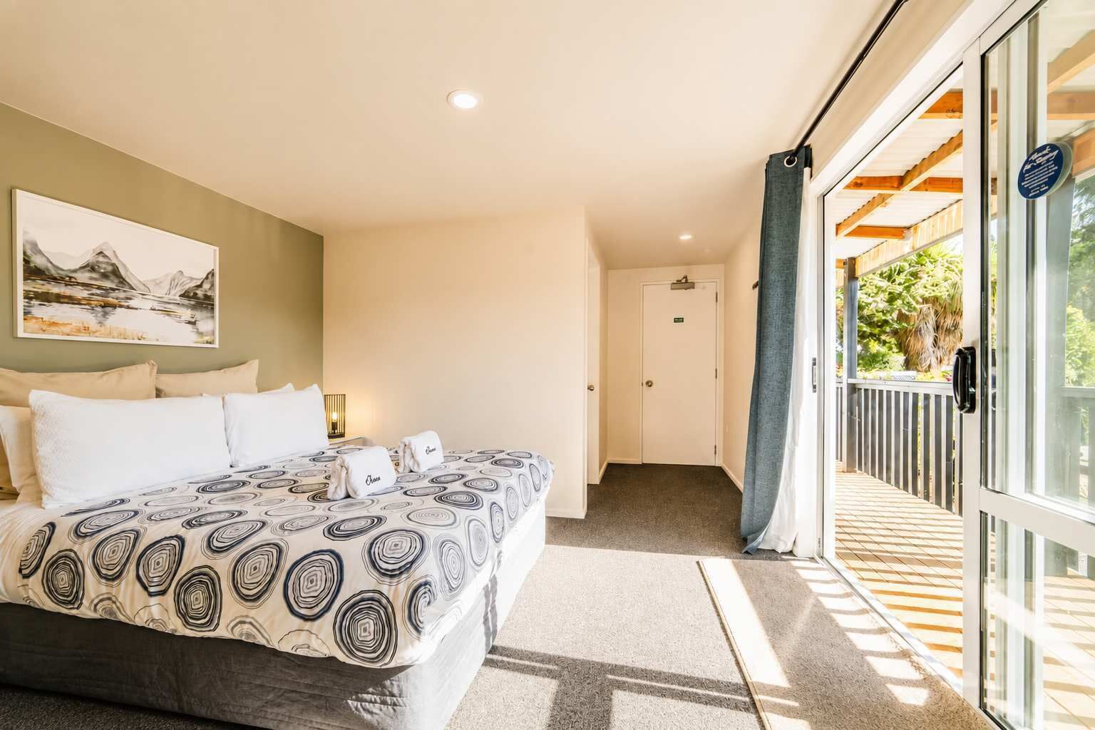 A hotel room with a bed, a painting on the wall, a door leading to a balcony, and a view of greenery outside.