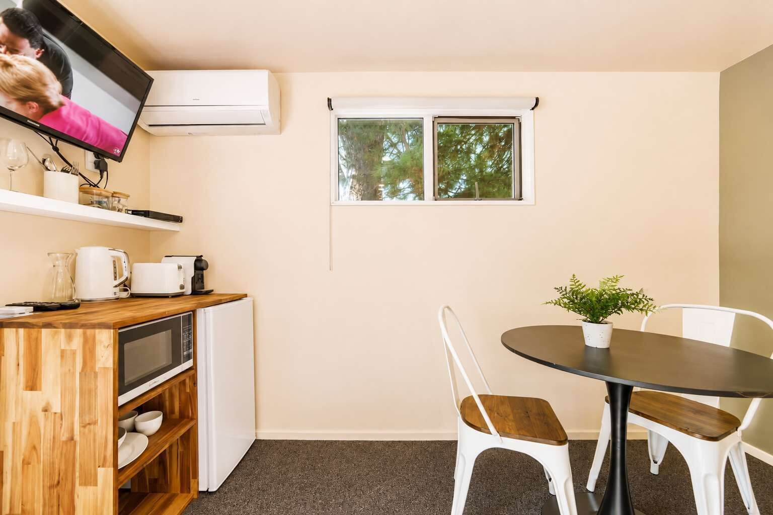 A compact kitchenette with a microwave, toaster, and coffee maker, a small dining table with chairs, and a window with a view of trees.