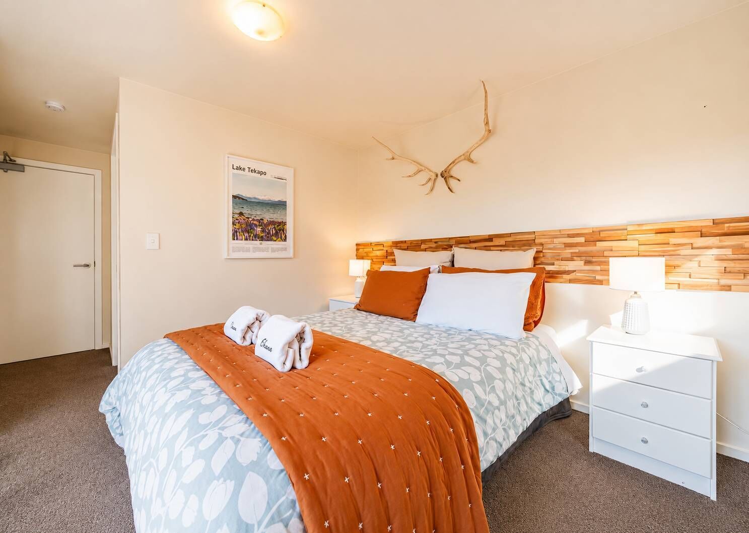 Una habitación de hotel con cama, mesita de noche, lámpara, un cartel de Lago Tekapo y cabecero de madera.