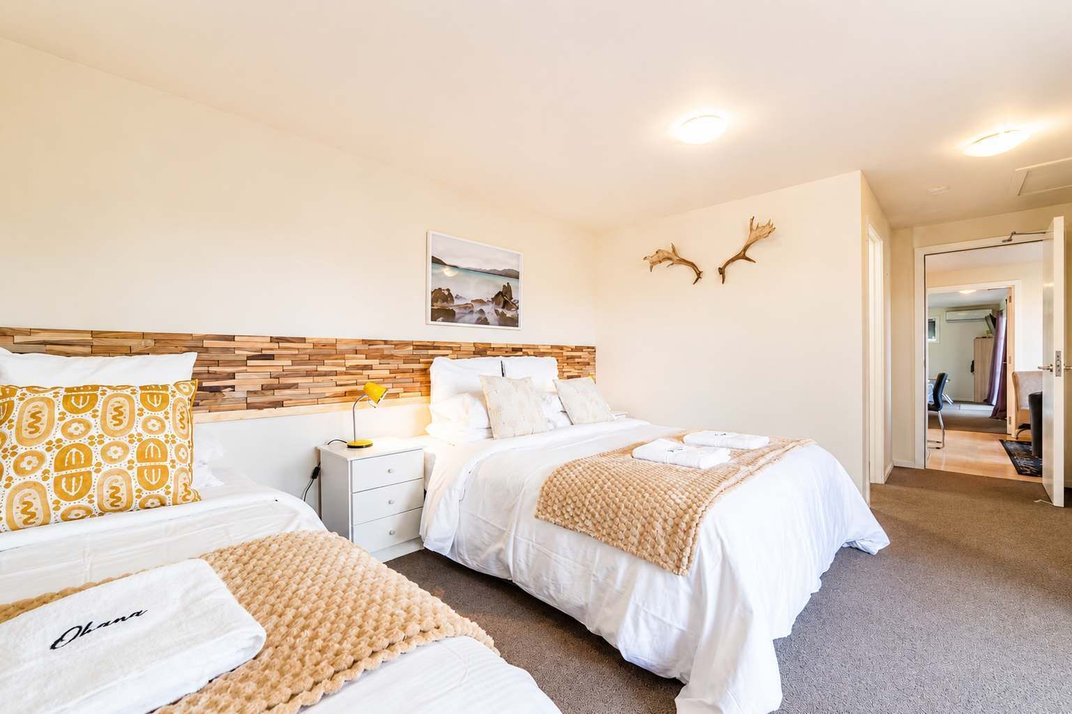 Ein Hotelzimmer mit zwei Betten, einem hölzernen Kopfteil, einem Nachttisch mit Lampe, einem gerahmten Bild an der Wand und einer Tür zu einem weiteren Raum.