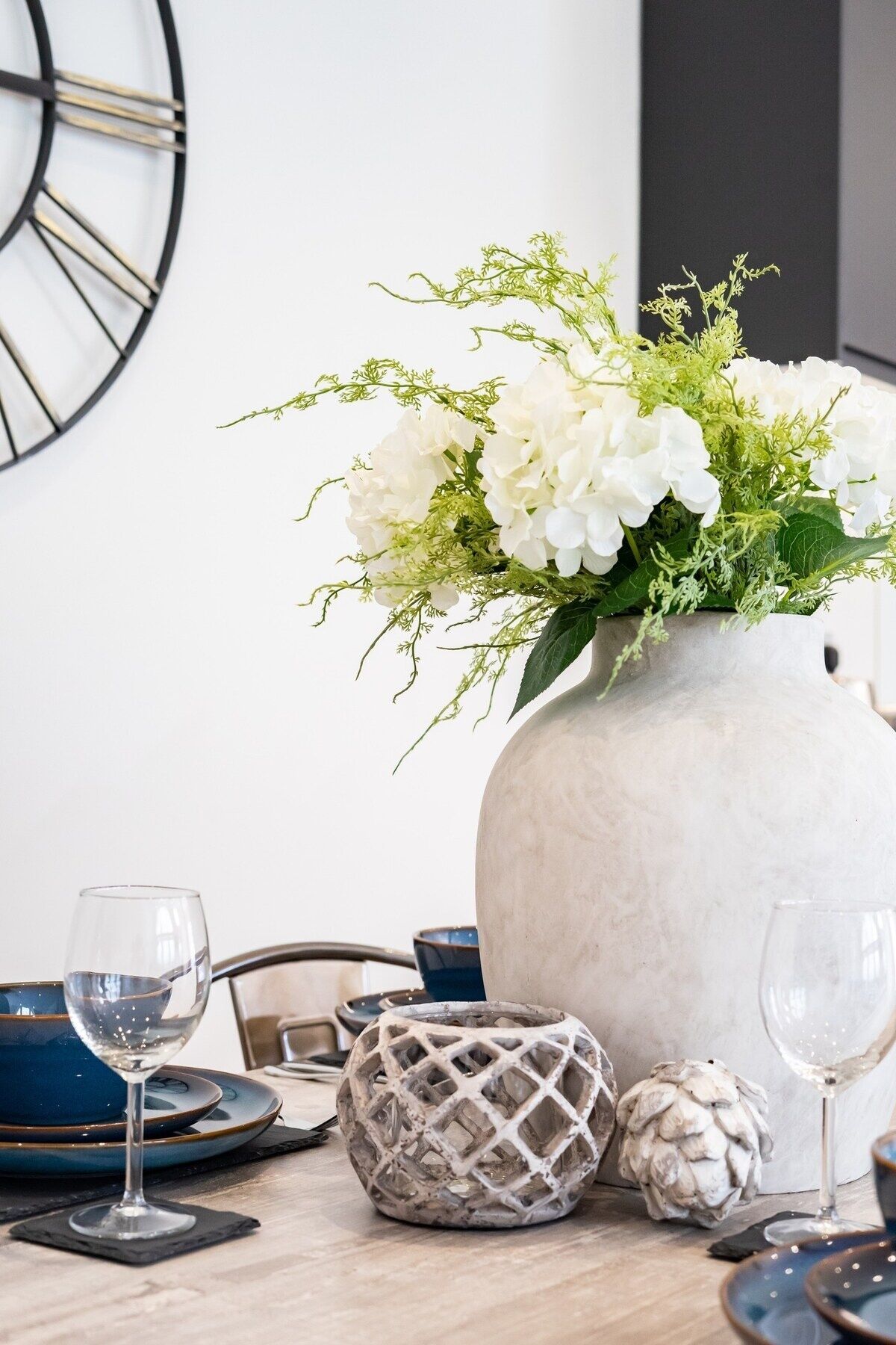 Een eettafel met een witte vaas vol witte bloemen en groen, blauwe borden en wijnglazen.