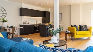 A modern living room with a sofa, a yellow armchair, and a coffee table. There is a kitchen area with dark cabinets and a small island. - The Old Glassworks by Bevolve - Free Parking (Shrewsbury)
